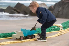 Mały chłopiec bawiący się na plaży zabawkową ciężarówką z piaskiem w ubraniu na chłodną pogodę