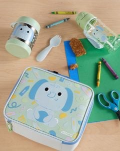 Torba termiczna na lunch z motywem niebieskiego słonia dla dziecka wraz z pojemnikiem i sztućcami na drewnianym stole. Skip Hop Torba Termiczna Lunch Bag Słoń Besities Squad
