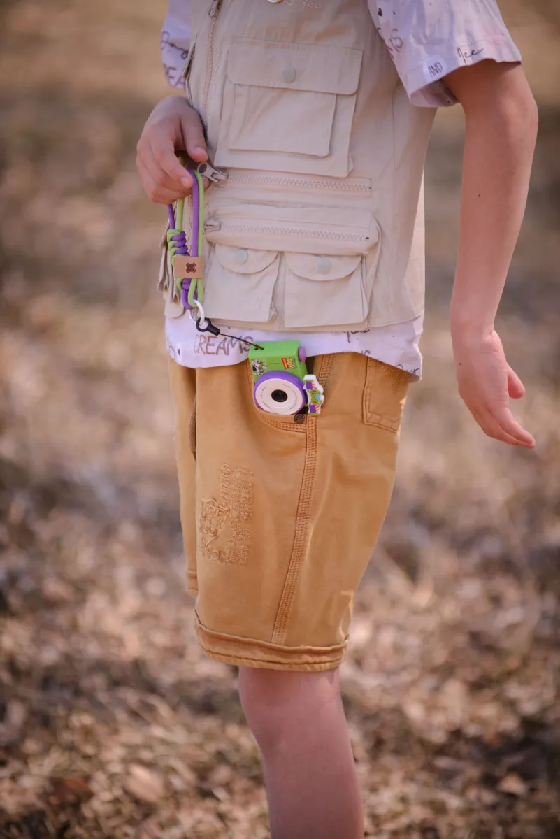 Mini aparat fotograficzny dla dzieci w kolorze zielonym z motywem Buzz Astral z Toy Story zawieszony na sznurku przy spodenkach chłopięcych beżowych. Kiimento Aparat Mini dla Dzieci Buzz Astral z Toy Story