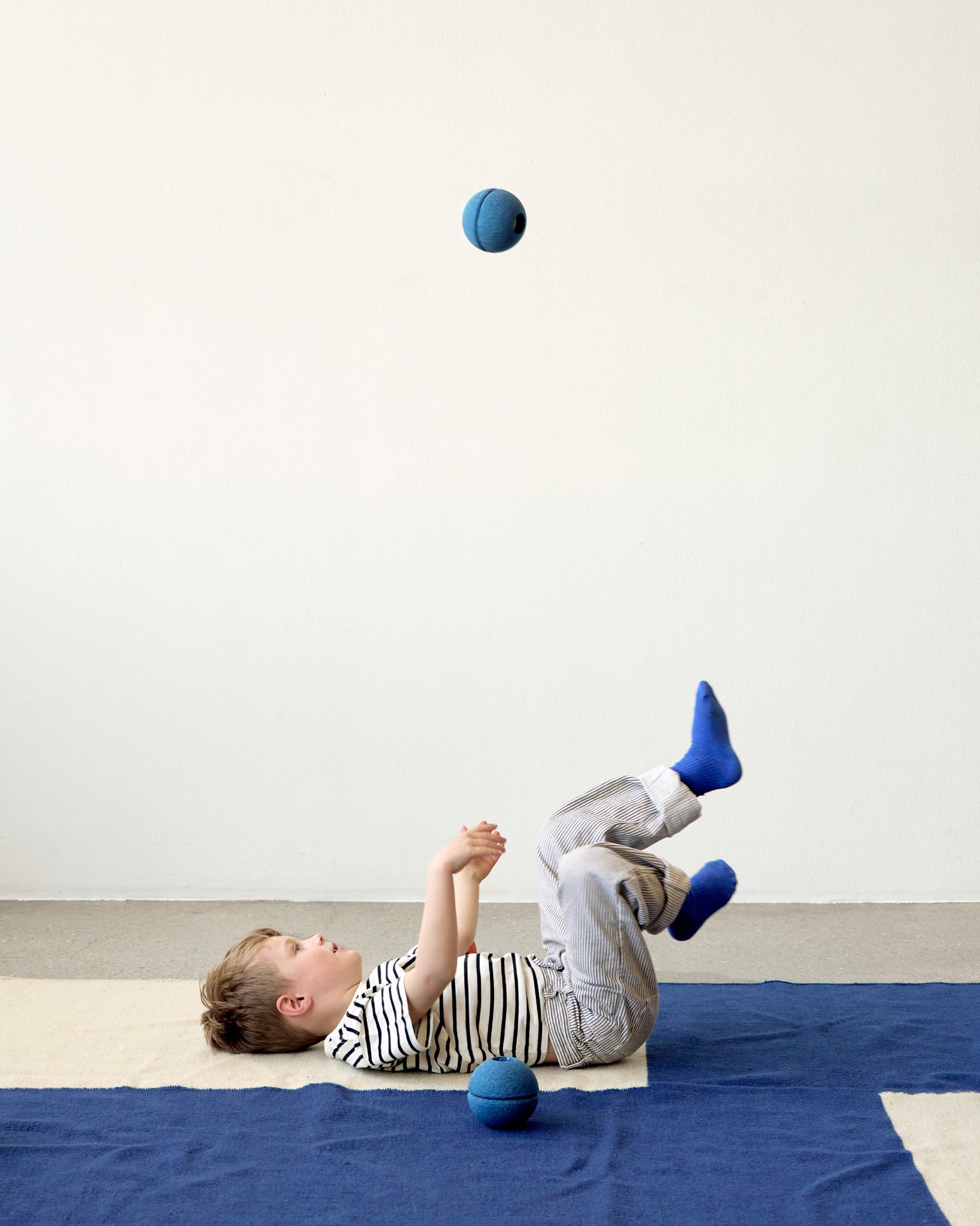 Dziecko leżące na plecach na niebieskim dywanie z piankową piłką w kolorze głęboki niebieski i jasny niebieski. MODU Foam Ball piłka piankowa deep blue / sky blue