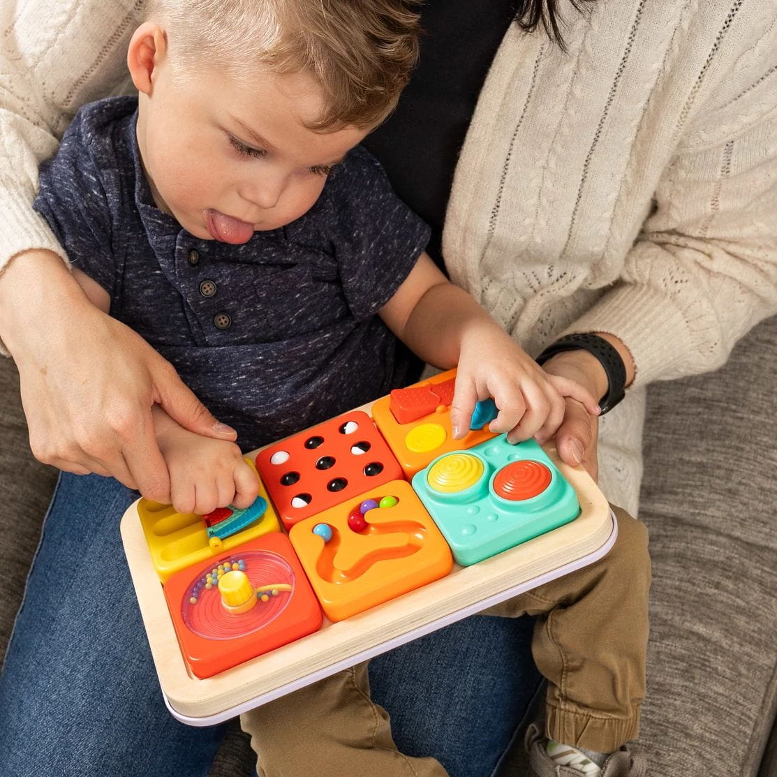 Dziecko bawiące się sensorycznym drewnianym zestawem z kolorowymi kwadratami i zgarniaczem kuleczek na kolanach osoby dorosłej. Fat Brain Toy Co. PlayTab Sensoryczne Kwadraty Zestaw 3 ze Zgarniaczem Kuleczek