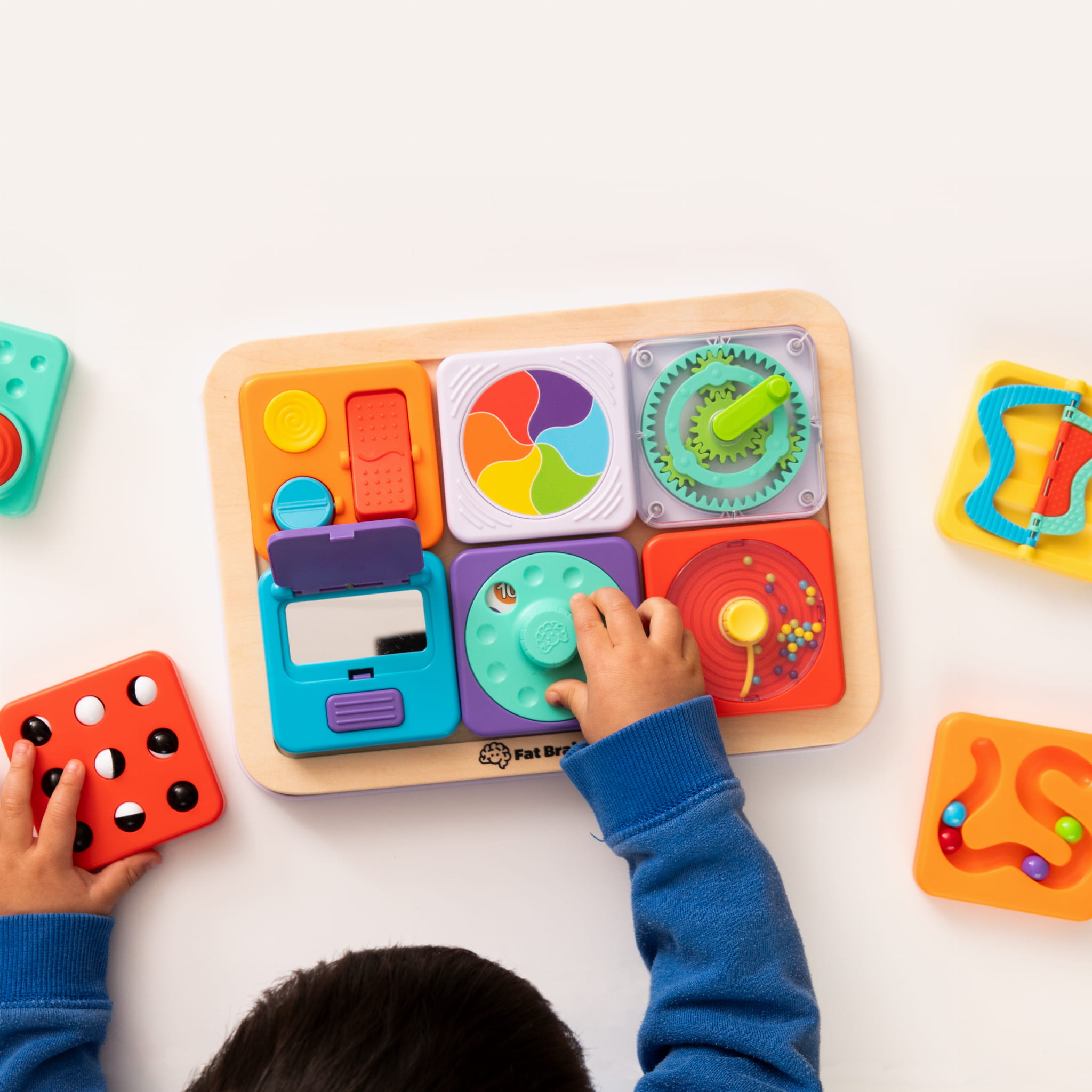 Dziecko bawiące się sensorycznym zestawem z kolorowymi kwadratami i elementami do przesuwania i obracania na drewnianej podstawce. Fat Brain Toy Co. PlayTab Sensoryczne Kwadraty Zestaw 3 ze Zgarniaczem Kuleczek