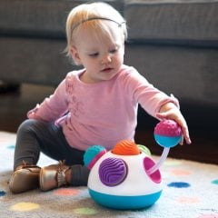 Niemowlę bawiące się sensoryczną zabawką z kolorowymi elementami do chwytania i obracania na dywanie. Fat Brain Toy Co. Sensoryczna Pracownia Klickity