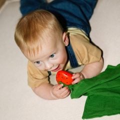 Niemowlę trzymające czerwony gryzak w kształcie truskawki z zielonym materiałem dla dziecka. Oli & Carol Przytulanka z Gryzakiem Big Doudou Truskawka Sweetie