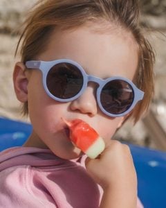 Dziecko w jasnoniebieskich okularach przeciwsłonecznych dla dzieci 1.5-3 lat jedzące loda. OKKY Okulary przeciwsłoneczne light blue 1.5-3 lat