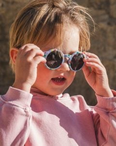 Dziecko w różowej bluzie zakłada niebieskie okulary przeciwsłoneczne z wzorem dla dzieci 1.5-3 lat. OKKY Okulary przeciwsłoneczne print blue 1.5-3 lat