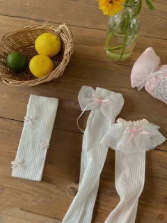 Białe podkolanówki dziecięce z kokardkami i koronką na drewnianym tle obok koszyka z cytrusami i kwiatów w wazonie. Fig&Pear Białe podkolanówki z kokardkami i koronką 3-pak