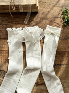 Trzy pary białych koronkowych podkolanówek dla dziecka z kokardkami i falbankami na drewnianym tle. Fig&Pear Koronkowe podkolanówki 3-pak