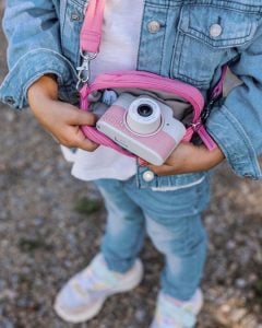 Różowy aparat dla dzieci na pasku z torbą na ramię trzymany przez dziecko w dżinsowej kurtce. Hoppstar Expert Blush Zestaw aparat dla dzieci i torba na aparat