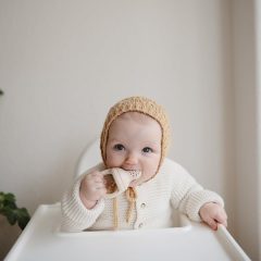 Dziecko w kremowym swetrze i czapce trzyma gryzak do podawania pokarmu przy krzesełku do karmienia. Mushie Gryzak do Podawania Pokarmu 2-pak Shifting Sand