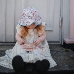 Dziecko w kwiecistym kapeluszu trzyma różową sensoryczną przeplatankę w kształcie kwiatka. Mushie Tablica sensoryczna labirynt przeplatanka Kwiatek
