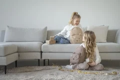 Dwie dziewczynki bawiące się na dywanie przy szarej sofie z plecakami w pokoju dziennym