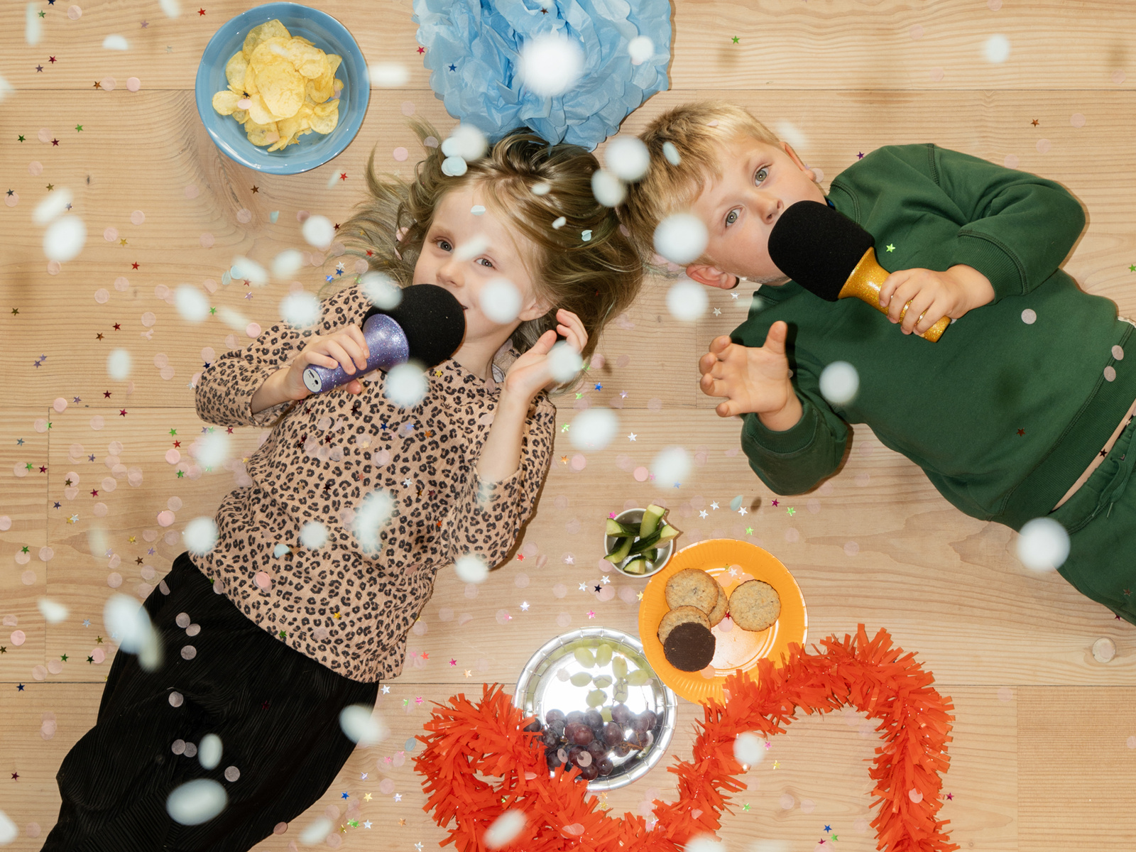 Dzieci śpiewające do mikrofonów karaoke w kolorze fioletowym i czarnym na podłodze, z konfetti i ozdobami wokół. Lalarma Mikrofon Karaoke Glitter Groove Fioletowy
