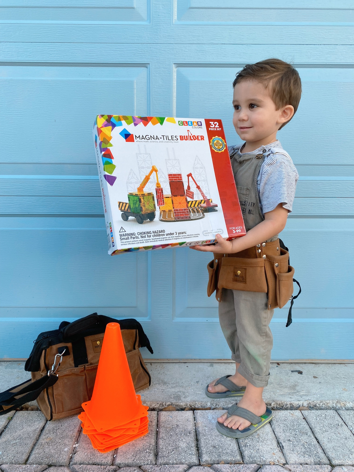 Dziecko trzyma pudełko z klockami magnetycznymi z kolorową budowlą i pomarańczowym stożkiem na ilustracji. Magna-Tiles Klocki Magnetyczne Builder 32el