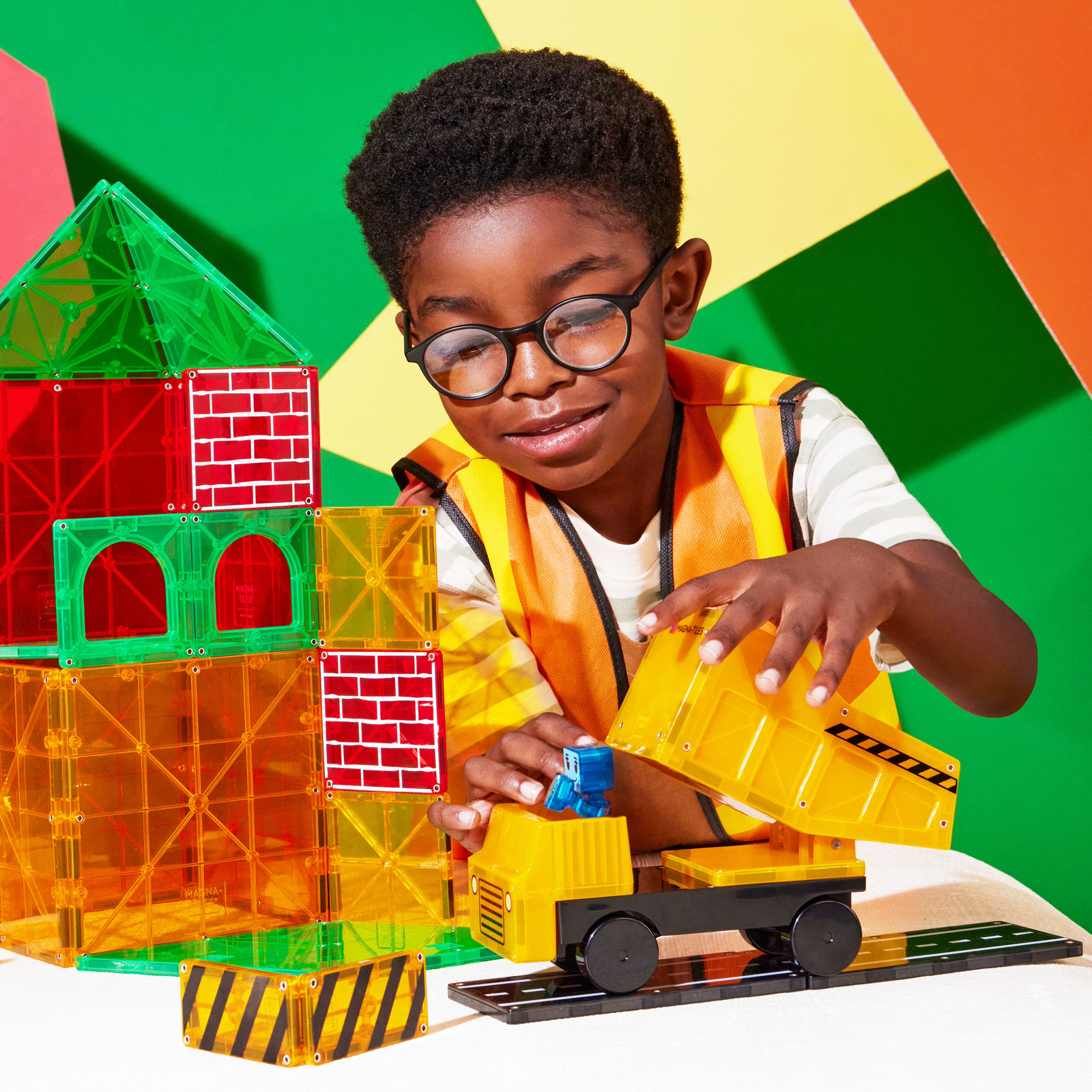 Dziecko bawi się kolorowymi klockami magnetycznymi i żółtą ciężarówką budowlaną przy wieży z przezroczystych elementów. Magna-Tiles Klocki Magnetyczne Builder XL 50el