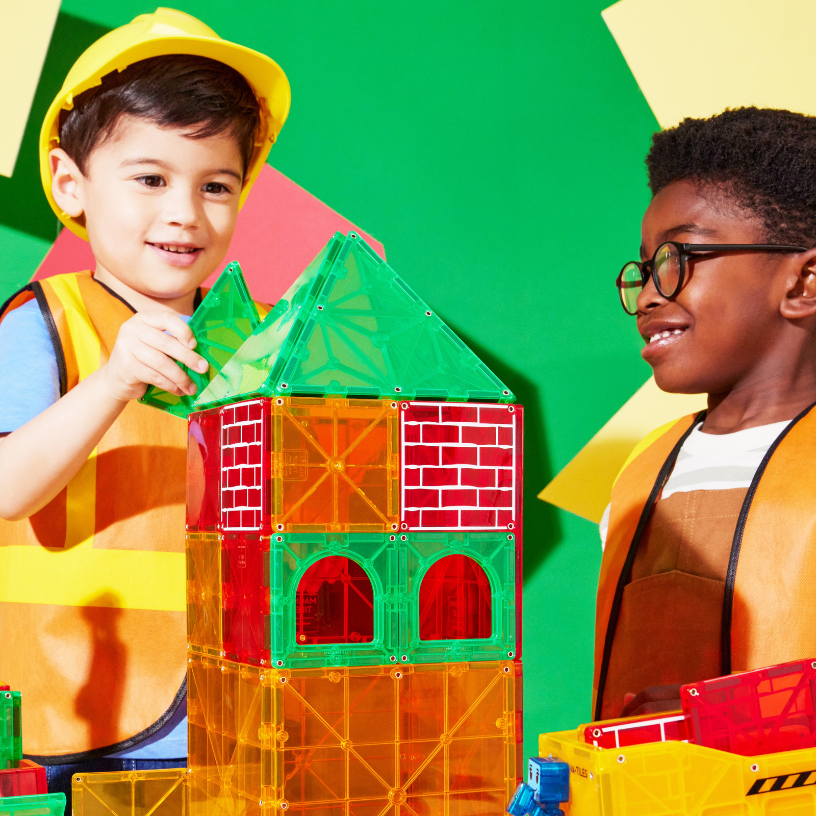 Dzieci bawiące się kolorowymi klockami magnetycznymi tworzącymi wieżę z zielonym dachem i czerwonymi ścianami. Magna-Tiles Klocki Magnetyczne Builder XL 50el