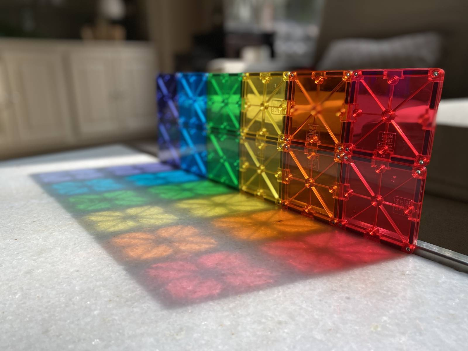 Kolorowe magnetyczne klocki z przezroczystego plastiku ustawione w rządku na podłodze, tworzące tęczowy wzór światła. Magna-Tiles Klocki Magnetyczne Classic 100el