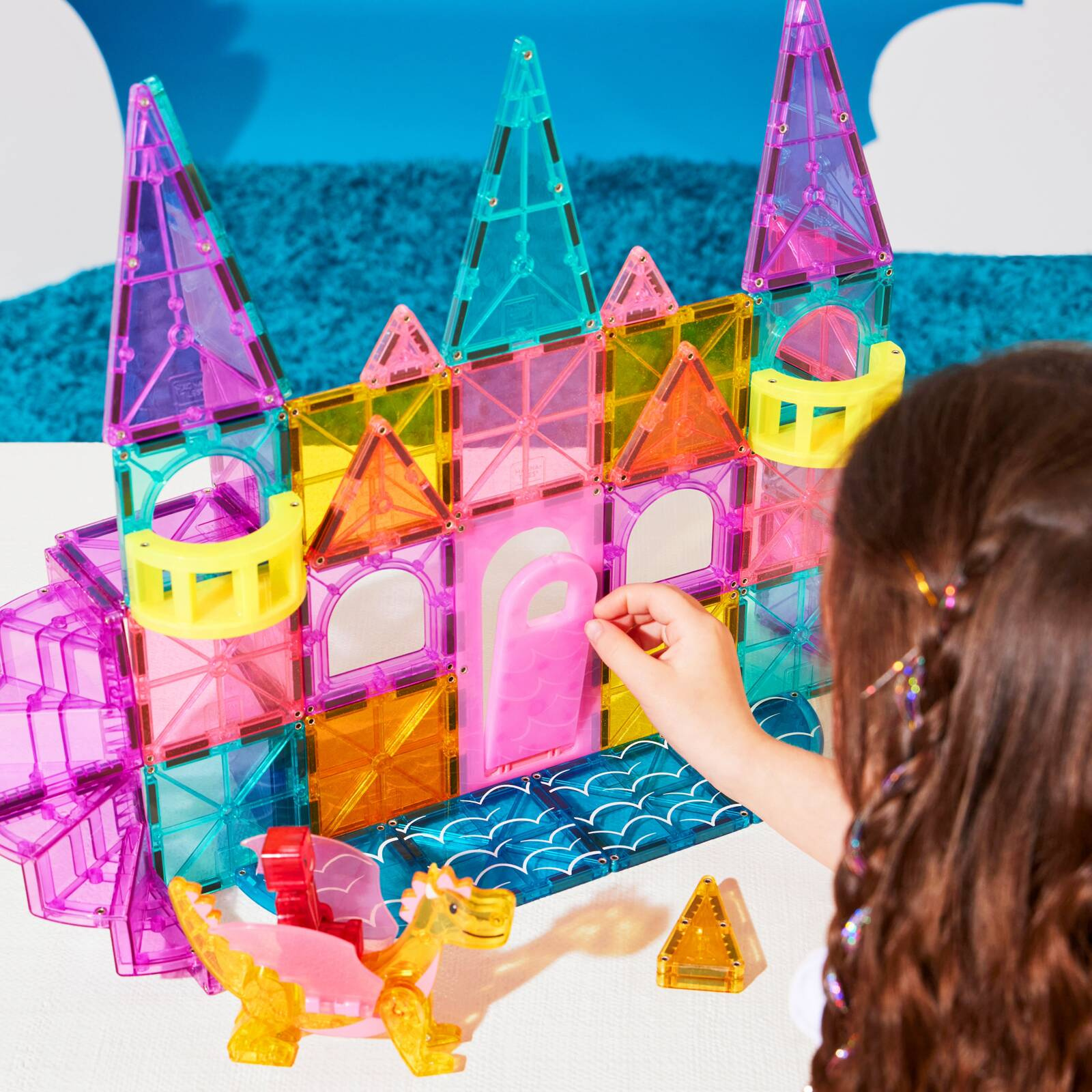 Kolorowy zamek z magnetycznych klocków z wieżyczkami i łukami, obok dziecko układa elementy. Magna-Tiles Klocki Magnetyczne Zamek DLX 48el