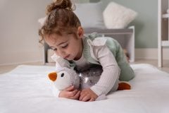Dziecko przytula białą pluszową zabawkę w kształcie pingwina z pomarańczowym dziobem na łóżku. Cloud B Projektor z pozytywką Pingwin ECO Tranquil Penguin