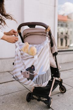Torba shopper w biało beżowe pasy z kolorowym wzorem, przypięta do rączki wózka dziecięcego, z pieczywem w środku